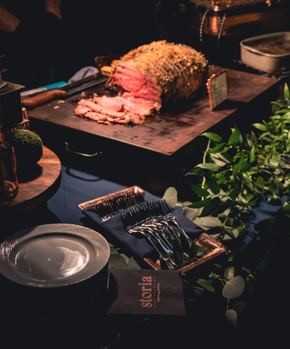 A partially cut up prime rib on a slab of wood. In the foreground sits a plate, a container full of silver forks, and a napkin with Storia's logo.